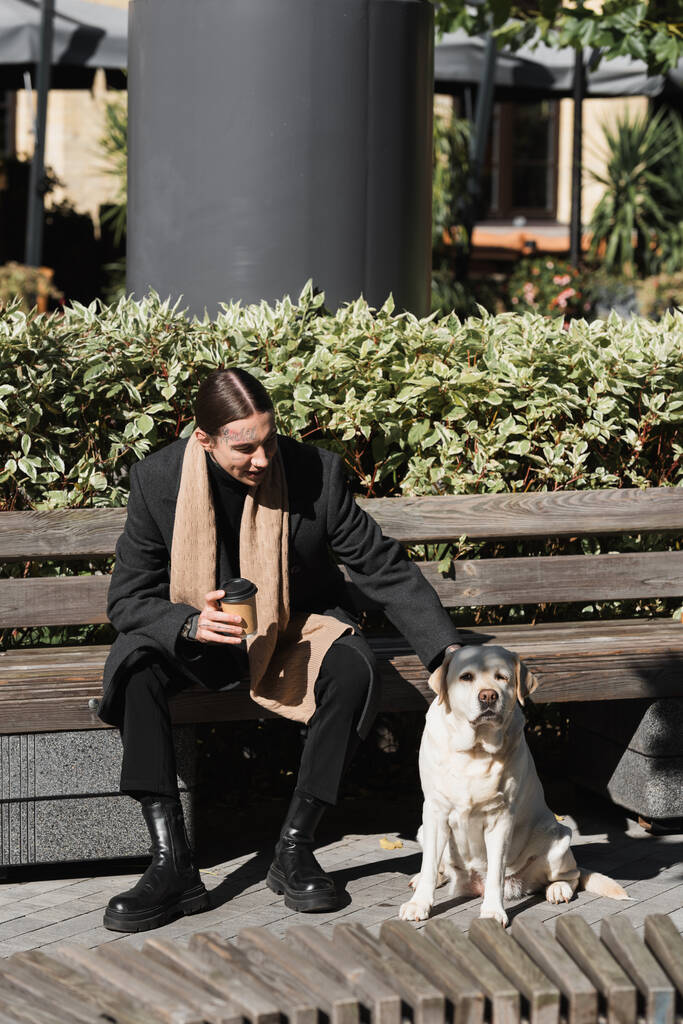 full length of happy tattooed man in coat holding takeaway drink and cuddling labrador  - Photo, Image