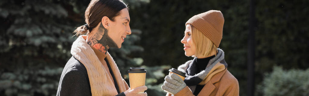 Seitenansicht einer fröhlichen Frau mit beigem Hut, die ihren tätowierten Freund im Mantel ansieht, während sie Coffee to go in der Hand hält, Banner  - Foto, Bild