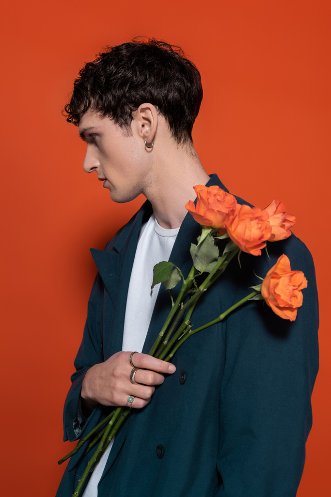 Side view of stylish young man in jacket holding orange roses for 8 march on red background  - Fotó, kép