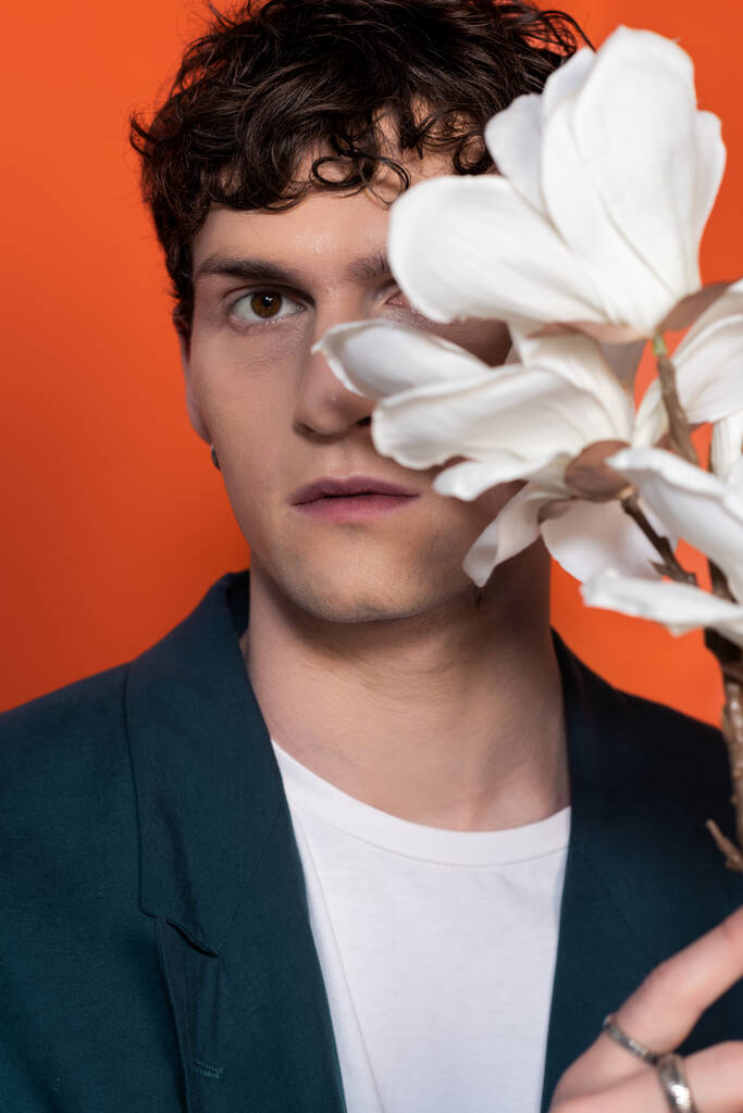 Curly man in jacket looking at camera near blurred magnolia flowers isolated on orange  - Photo, Image