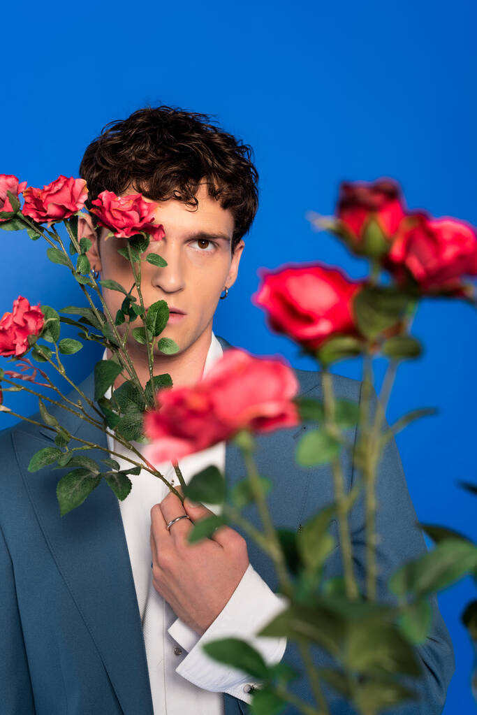 Curly man in stylish outfit covering face with flowers isolated on blue  - Photo, image