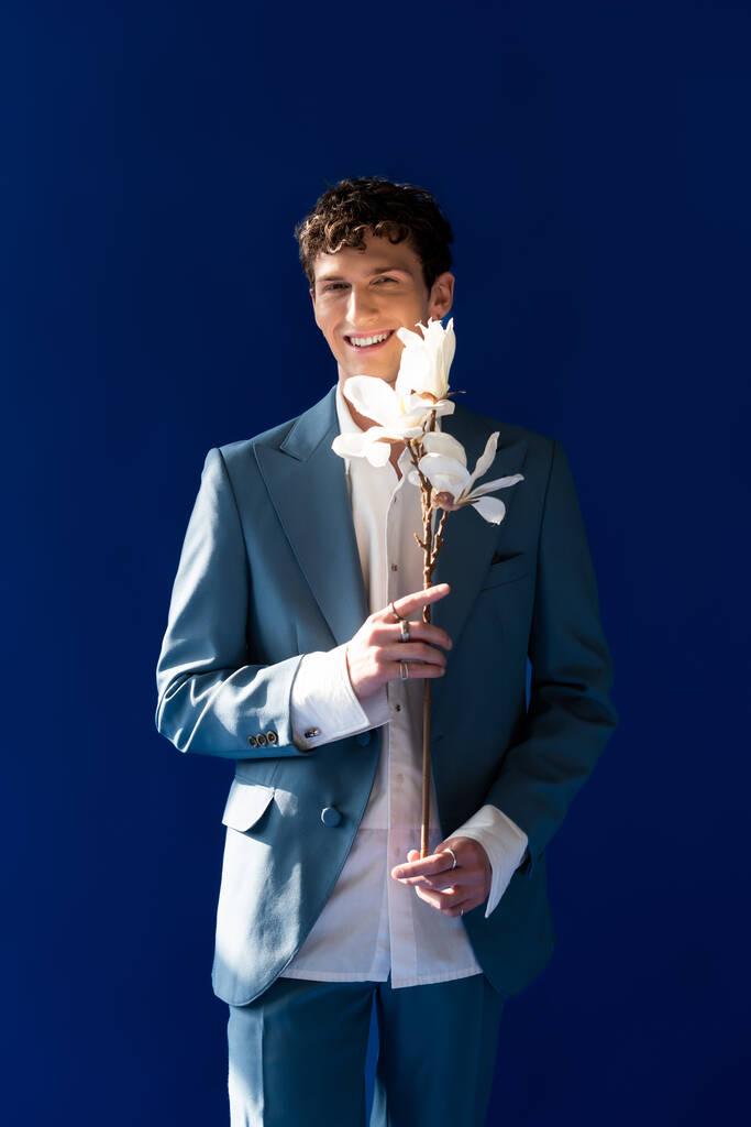 Стильный мужчина в костюме и рубашке, держащий ветку магнолии изолированной на синий цвет  - Фото, изображение