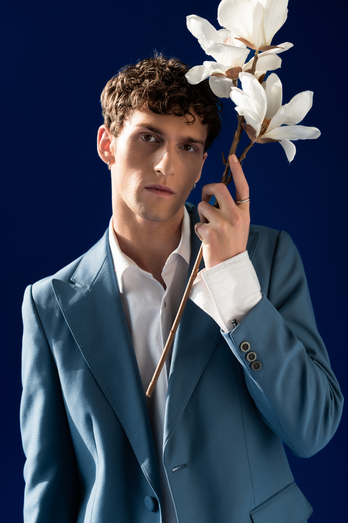 Portrait of stylish young man in jacket holding magnolia flowers isolated on navy blue  - Photo, Image