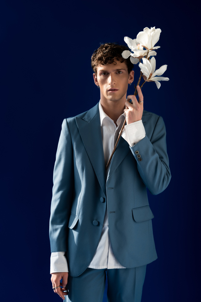 Модний молодий чоловік у костюмі тримає гілку магнолії ізольовано на синьому
  - Фото, зображення