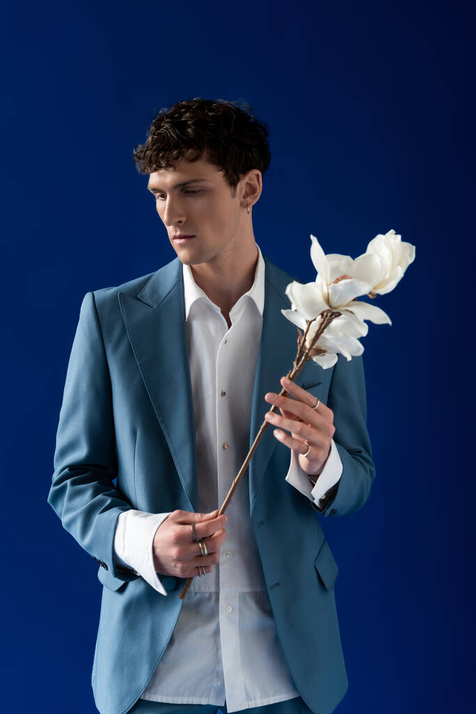 Hombre morena en chaqueta elegante y camisa que sostiene magnolia rama aislado en azul marino  - Foto, imagen