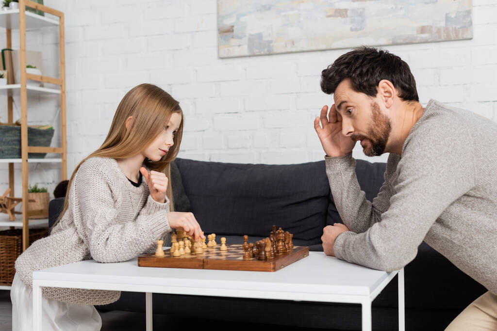 thoughtful man touching head while playing chess with preteen daughter in living room - Фото, изображение