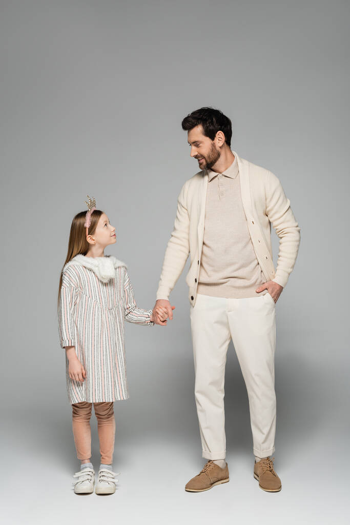 full length of happy man holding hands of kid in crown and standing with hand in pocket on grey - Foto, Imagen