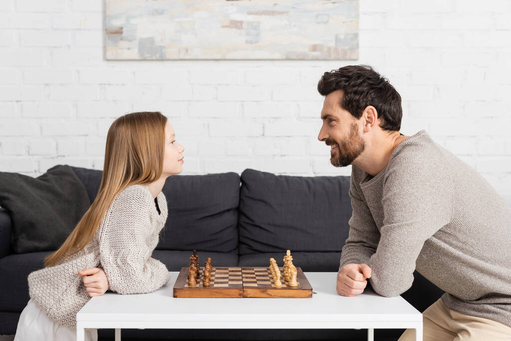 vista lateral da menina inteligente jogando xadrez com pai feliz na sala de estar  - Foto, Imagem