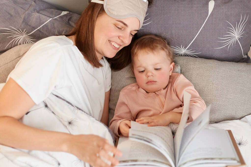 Εσωτερική λήψη χαμογελαστή όμορφη νεαρή μητέρα με την κόρη της διαβάζοντας το βιβλίο, ενώ στο κρεβάτι, ελκυστική γυναίκα ξοδεύει τεμπέλικο πρωινό με το μωρό της. - Φωτογραφία, εικόνα