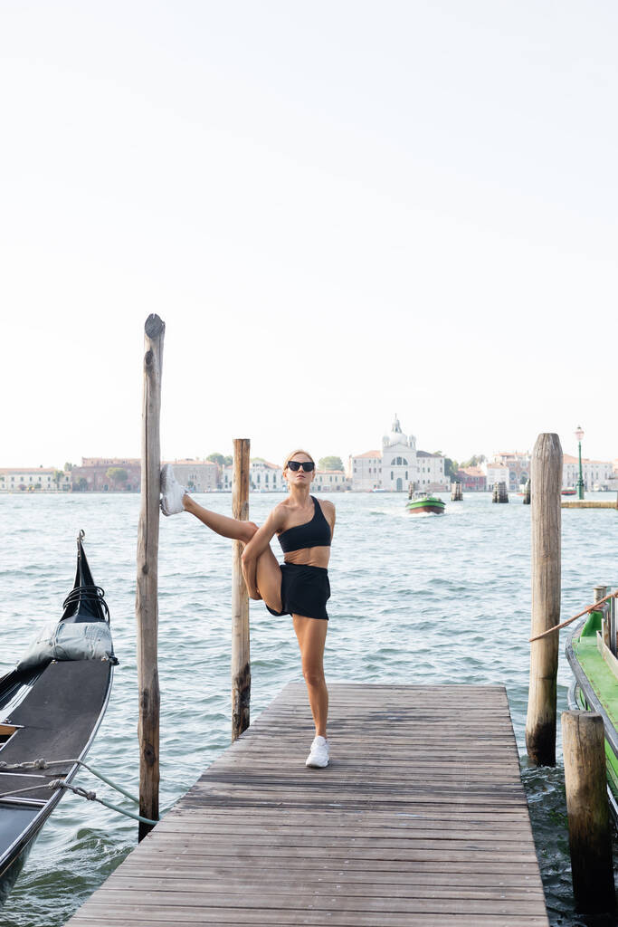 Flexible Sportlerin mit Sonnenbrille, weißen Turnschuhen, schwarzem bauchfreien Oberteil und kurzen Hosen beim Training auf einem Pier in Venedig  - Foto, Bild