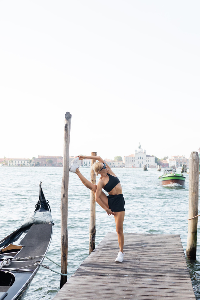 Güneş gözlüklü sarışın sporcu, beyaz spor ayakkabılı, siyah tişörtlü ve Venedik 'teki iskelede kısa şortlu.  - Fotoğraf, Görsel