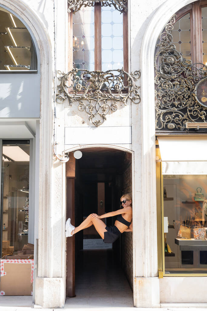 Smiling sportswoman in white sneakers, sunglasses, black crop top and shorts posing in arch of antique building in Venice  - Photo, Image
