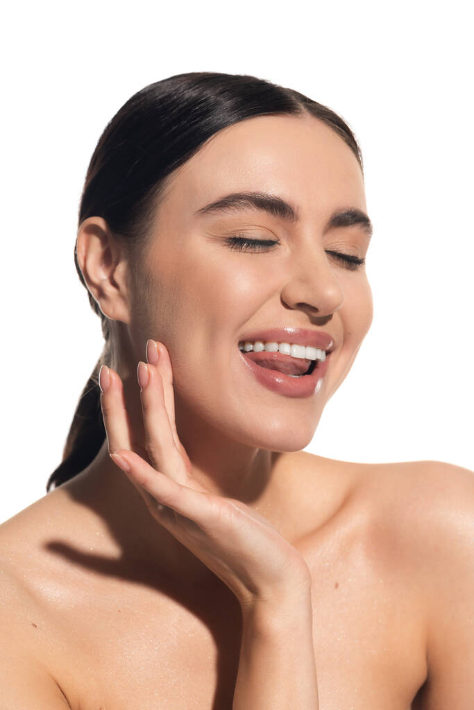 веселая молодая женщина с натуральным макияжем и обнаженным плечом касаясь щеки, торчащий язык изолирован на белом - Фото, изображение