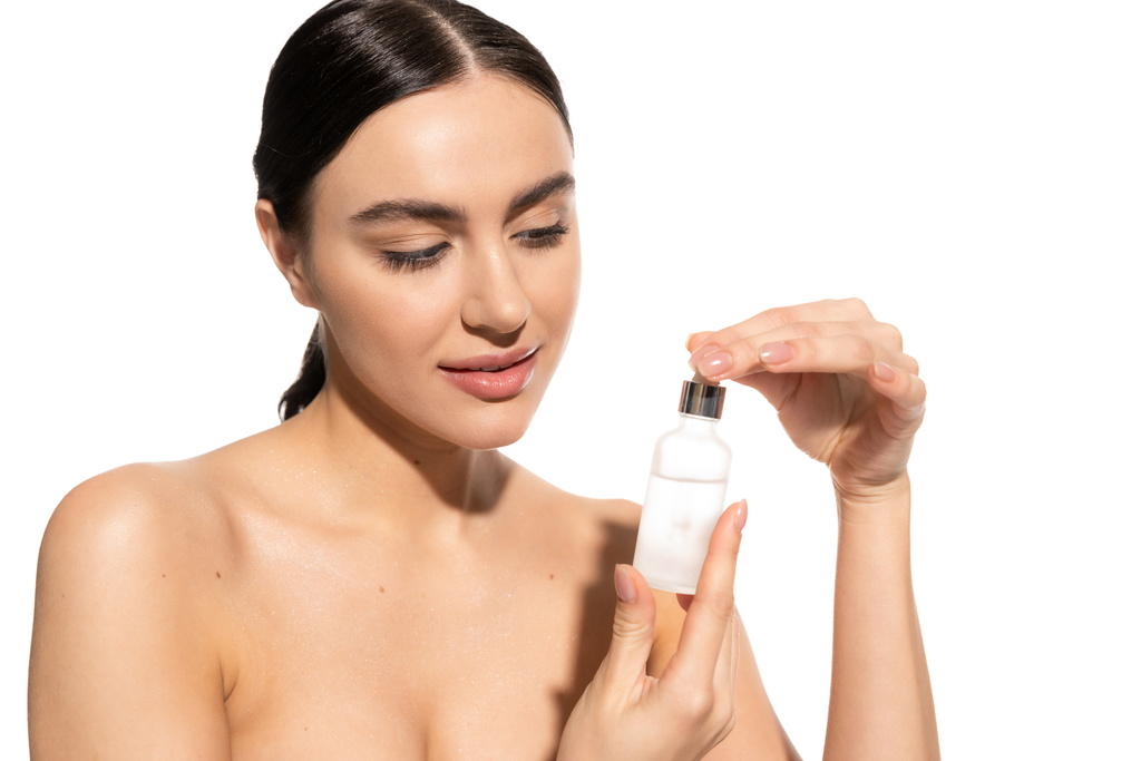 brunette woman with bare shoulders looking at bottle with moisturizing serum isolated on white  - Foto, Imagem