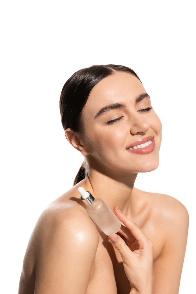 happy young woman with bare shoulders and closed eyes holding bottle with moisturizing serum isolated on white  - Fotografie, Obrázek