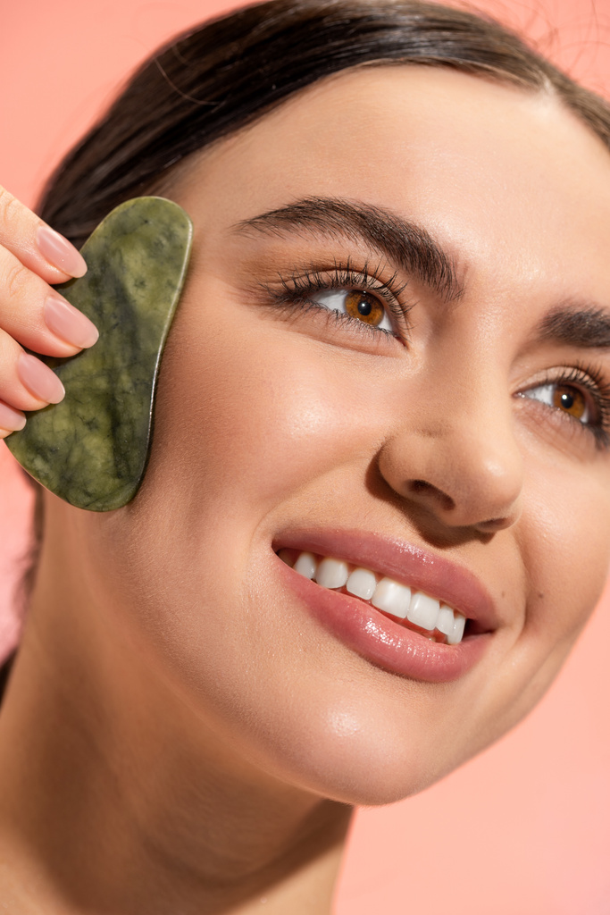 close up of happy woman with soft skin doing face massage with jade scraper isolated on pink  - Foto, afbeelding