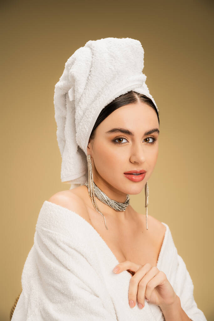 молодая женщина в ювелирных украшениях с белым полотенцем на голове, глядя на камеру на бежевом фоне  - Фото, изображение