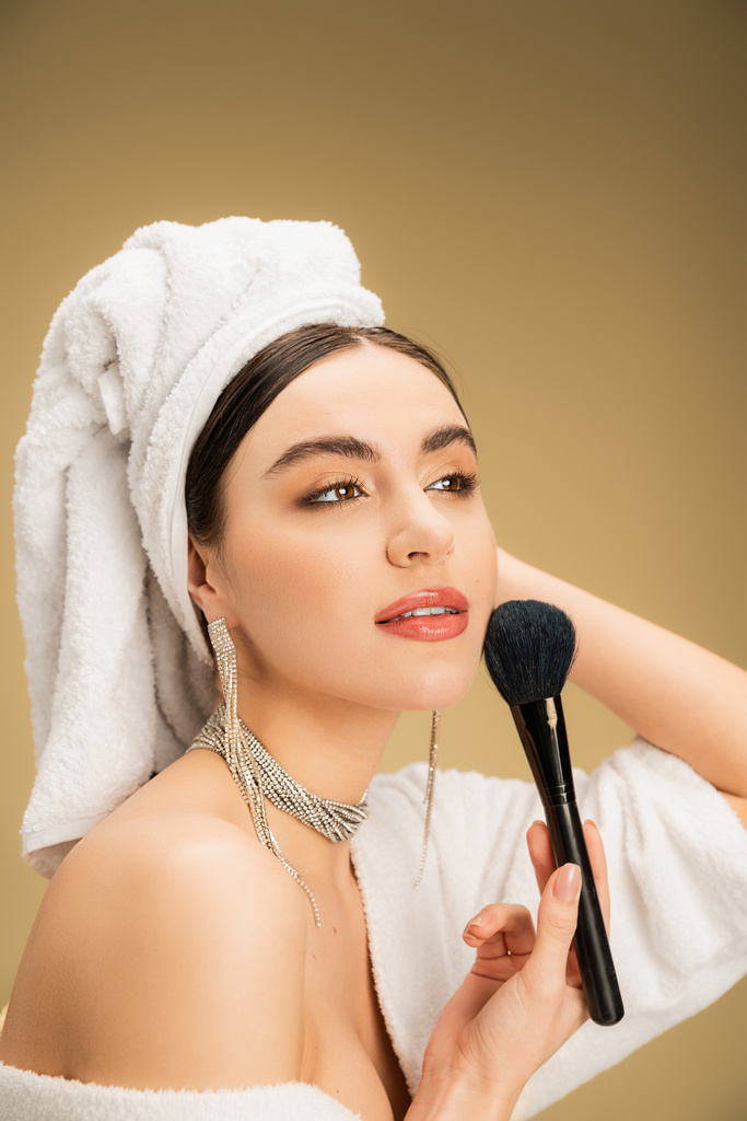 Başında beyaz havlu olan çekici kadın makyaj fırçasıyla bej arka planda yüz pudrası uyguluyor.  - Fotoğraf, Görsel