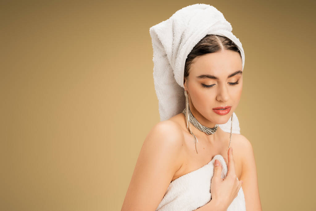 okouzlující mladá žena v šperky drží bílý ručník na béžové pozadí  - Fotografie, Obrázek