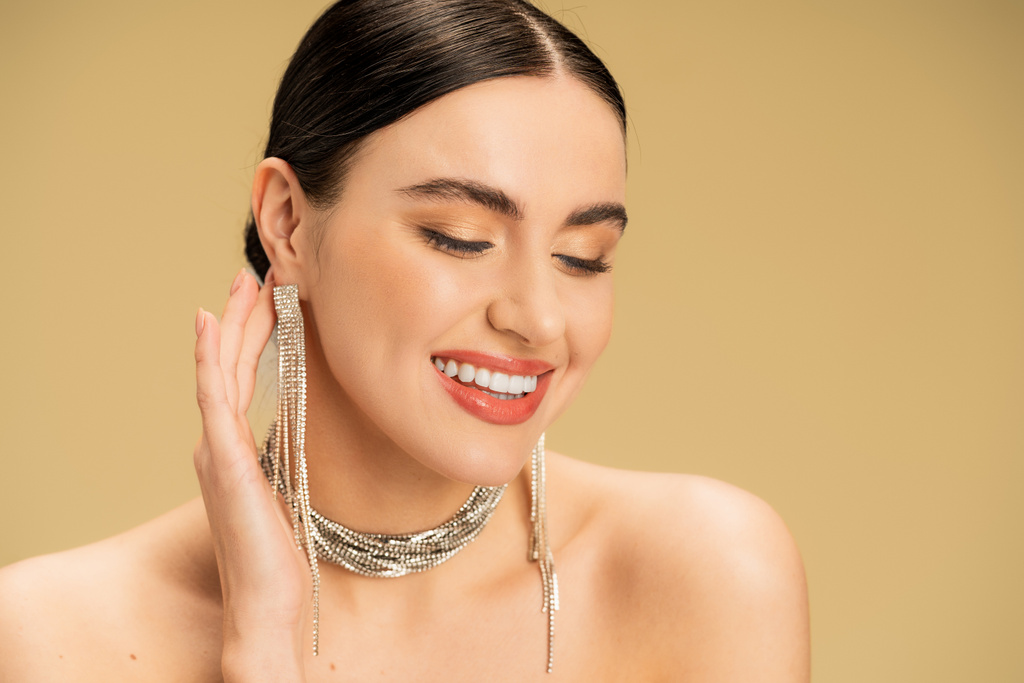 brunette young woman in necklace and earrings smiling isolated on beige  - Photo, Image