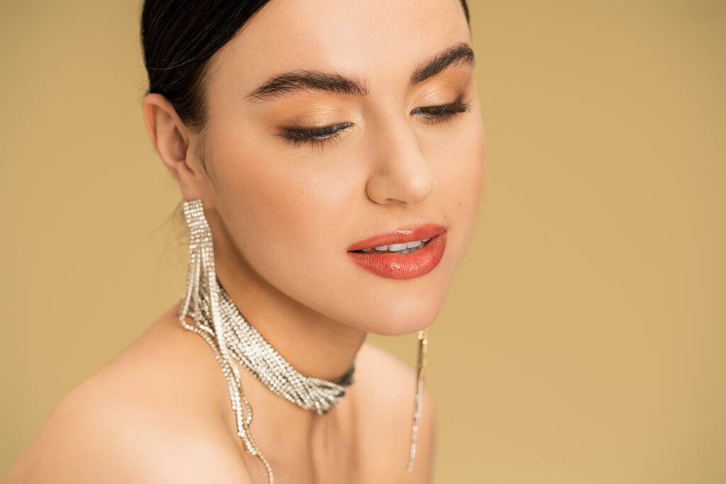 brunette woman in necklace and earrings looking away while posing isolated on beige  - Fotografie, Obrázek