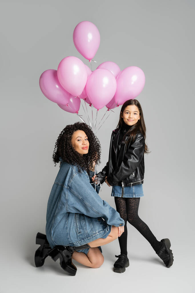 Fashionable woman looking at camera near daughter and pink balloons on grey background  - Photo, Image