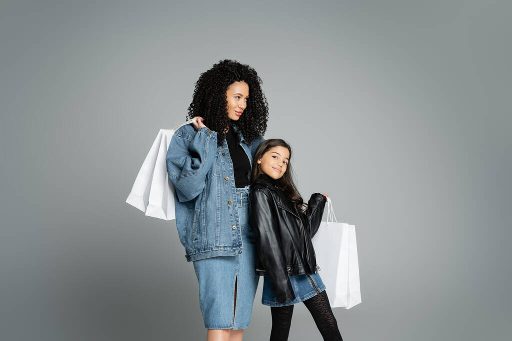 Trendy mother and daughter holding white shopping bags isolated on grey  - 写真・画像