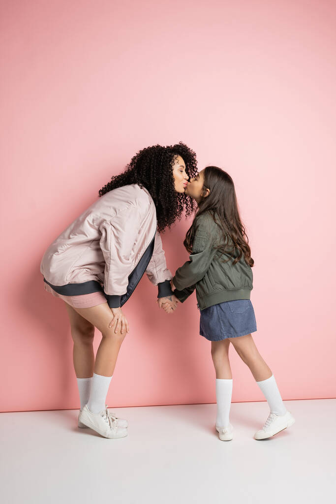 Side view of curly woman kissing stylish daughter on pink background  - Фото, изображение