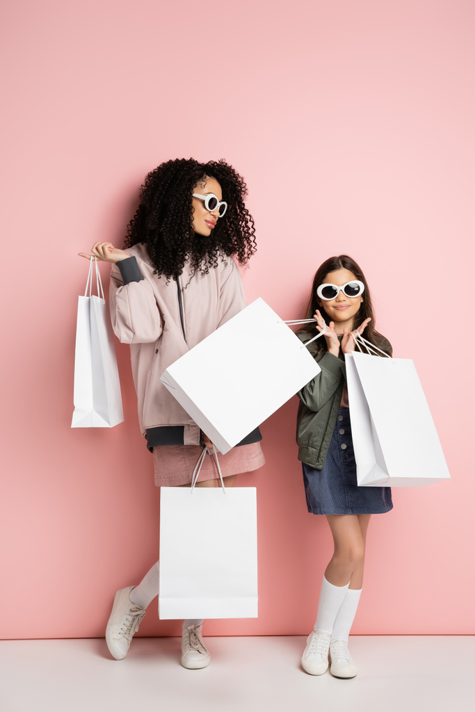 Stylish preteen girl holding purchases near mom in sunglasses on pink background  - Foto, imagen