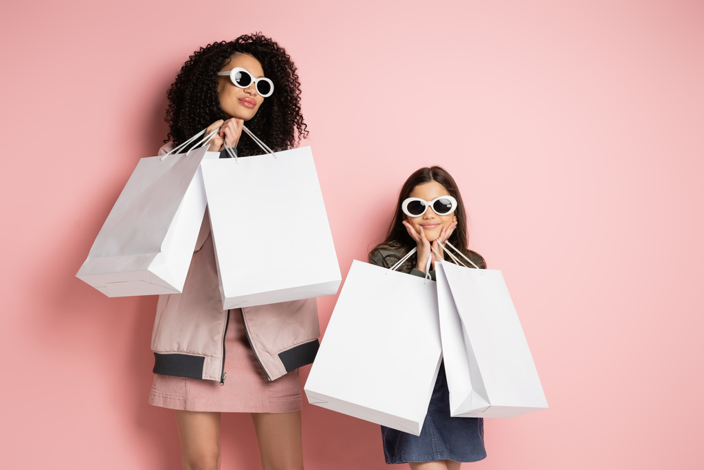 Fashionable mother and daughter in sunglasses holding shopping bags on pink background  - Fotografie, Obrázek