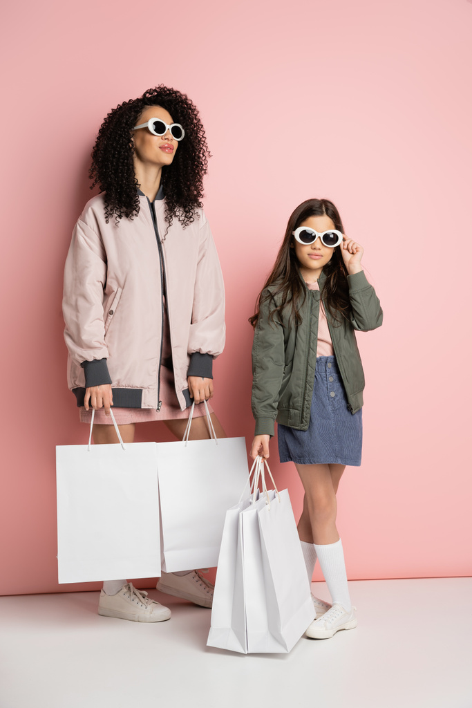 Fashionable mother and kid in sunglasses holding shopping bags on pink background  - Valokuva, kuva