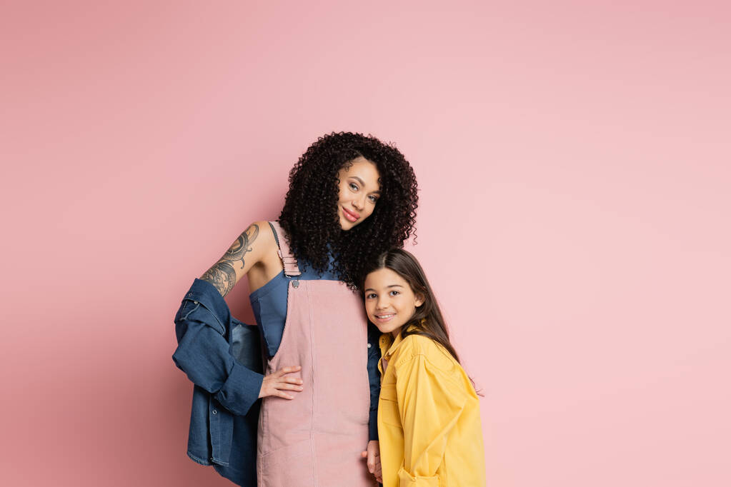 Smiling tattooed mother posing near daughter on pink background  - Foto, imagen