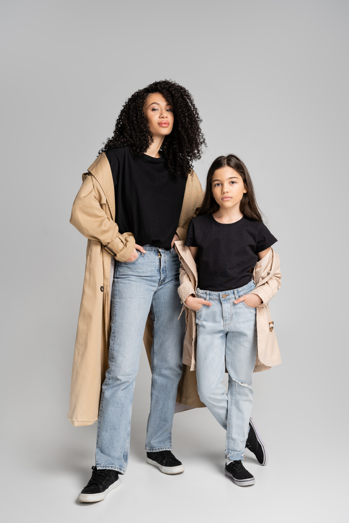 Curly mother in trench coat posing near stylish daughter on grey background  - Foto, immagini