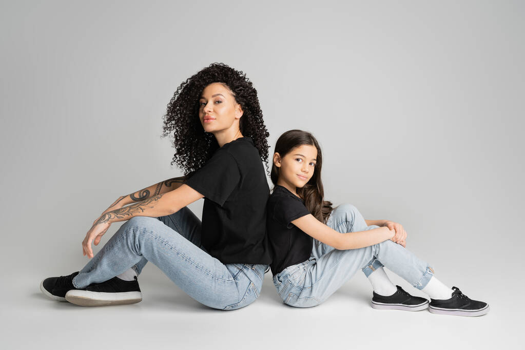 Tattooed mother and kid in jeans and t-shirts sitting back to back on grey background  - Fotó, kép