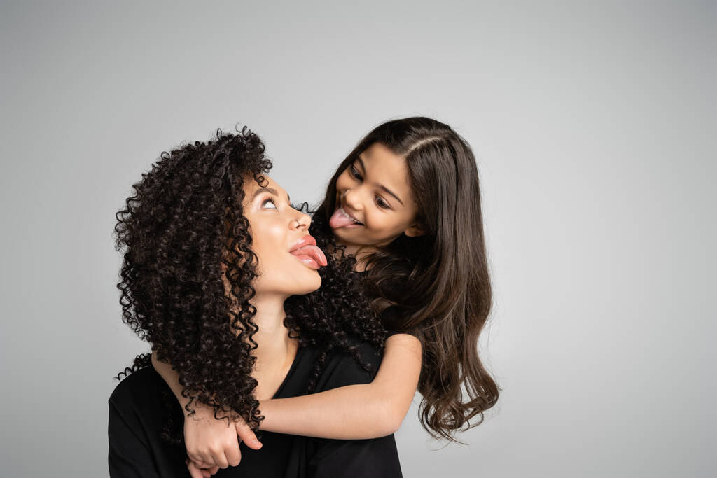 Preteen girl hugging mother and sticking out tongue isolated on grey  - Foto, imagen