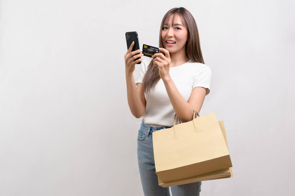 Μια νεαρή Ασιάτισσα που κρατάει τσάντα για ψώνια και πιστωτική κάρτα πάνω από το στούντιο λευκού φόντου, για ψώνια και χρηματοδότηση. - Φωτογραφία, εικόνα