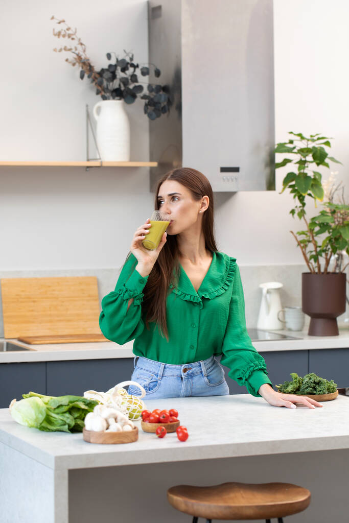 Een aantrekkelijke brunette drinkt een groene smoothie in haar keuken. Het concept van gezonde voeding, detox. Veganistische recepten. - Foto, afbeelding