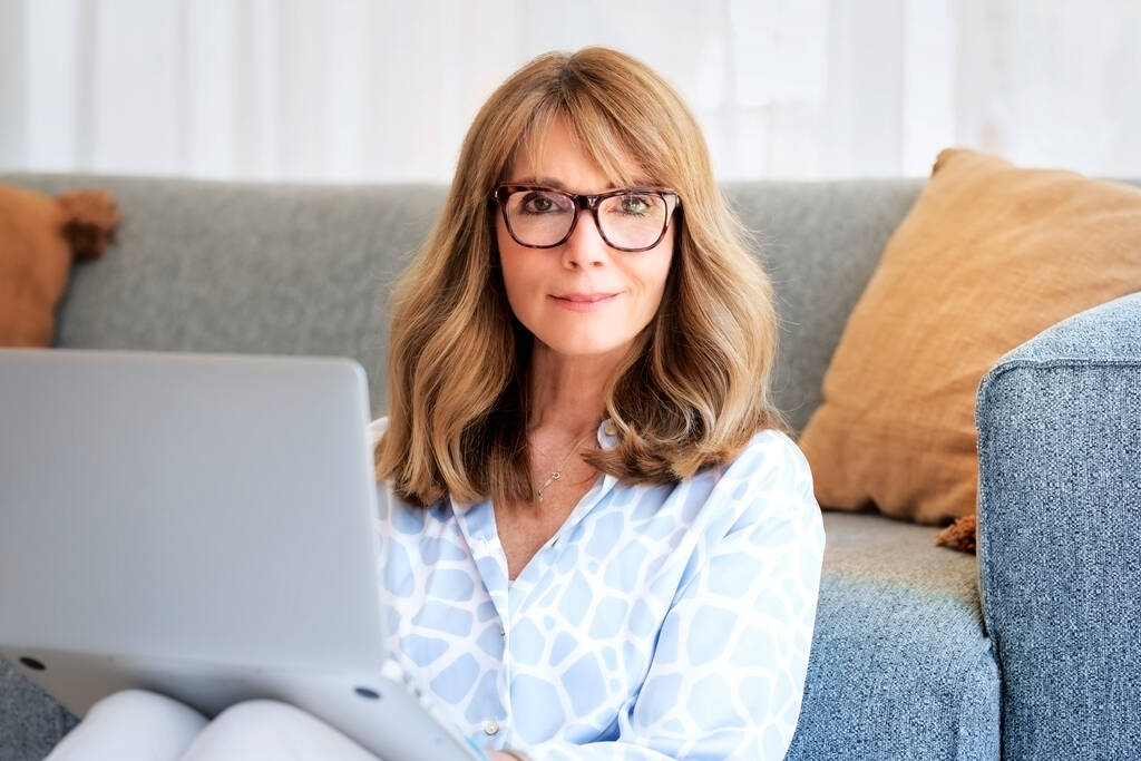 Közelkép egy középkorú nőről, aki otthon ül és laptopot használ a munkájához. Szőke hajú üzletasszony alkalmi ruhát és szemüveget visel, miközben otthon dolgozik. Belügyminisztérium.  - Fotó, kép