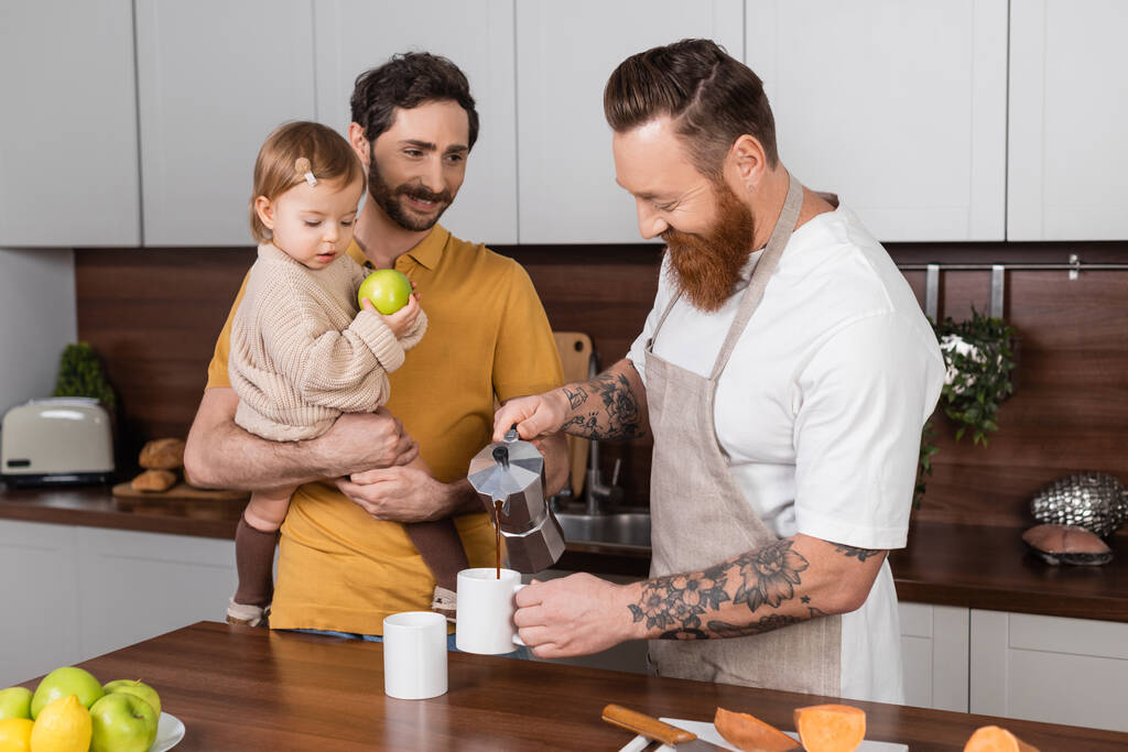 Sakallı eşcinsel adam, kocasının yanına kahve koyuyor mutfakta yeni yürümeye başlayan kızını tutuyor.  - Fotoğraf, Görsel