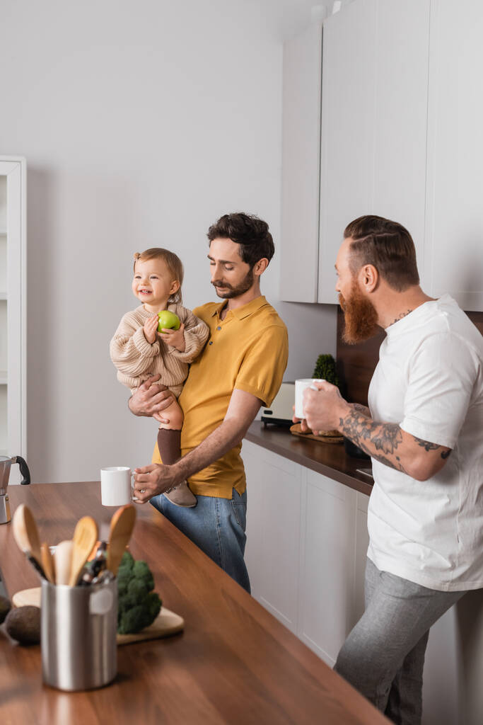 Gay couple holding coffee cups near smiling daughter with apple in kitchen  - Zdjęcie, obraz
