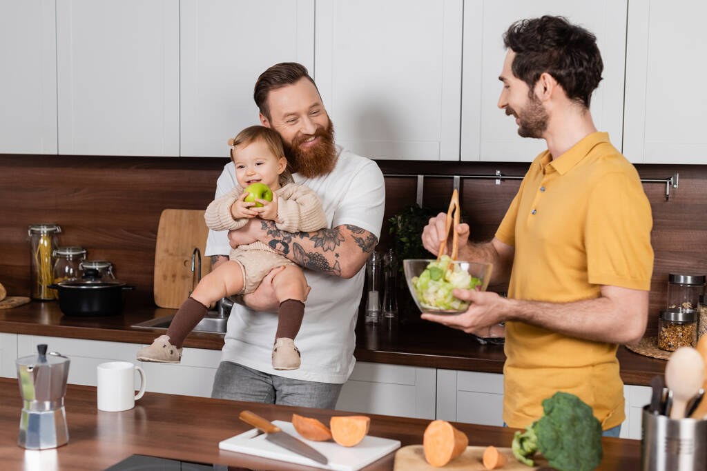 Lächelnder schwuler Mann umarmt Tochter mit Apfel, während Partner Salat in Küche kocht  - Foto, Bild