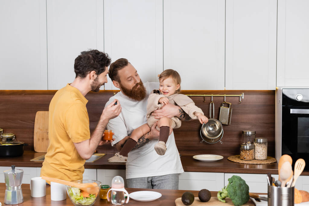 Gay parents holding baby food near displeased daughter in kitchen  - Photo, Image