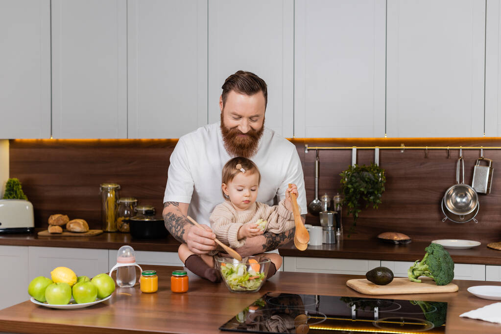 Tattooed father smiling and mixing salad near toddler daughter in kitchen  - Foto, imagen