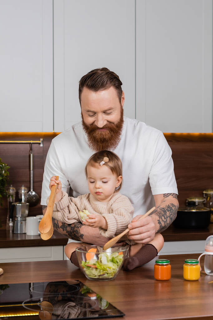 Smiling father cooking salad near daughter and baby food in kitchen  - Foto, Imagen