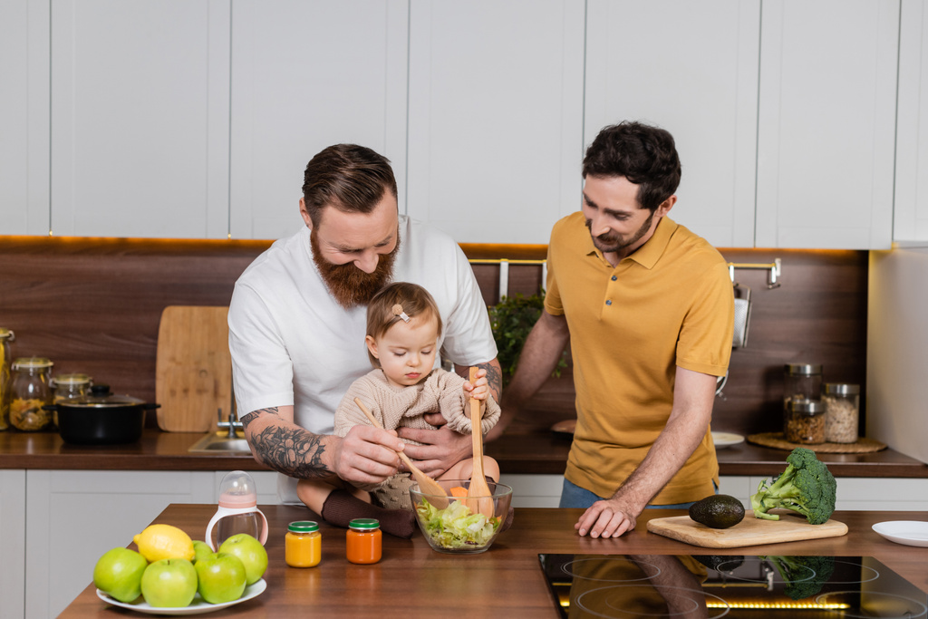 Gay man cooking salad near toddler daughter and partner in kitchen  - Foto, afbeelding