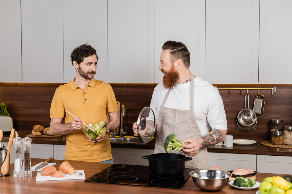 Homosexual man holding salad near partner cooking broccoli in kitchen  - Foto, Imagem