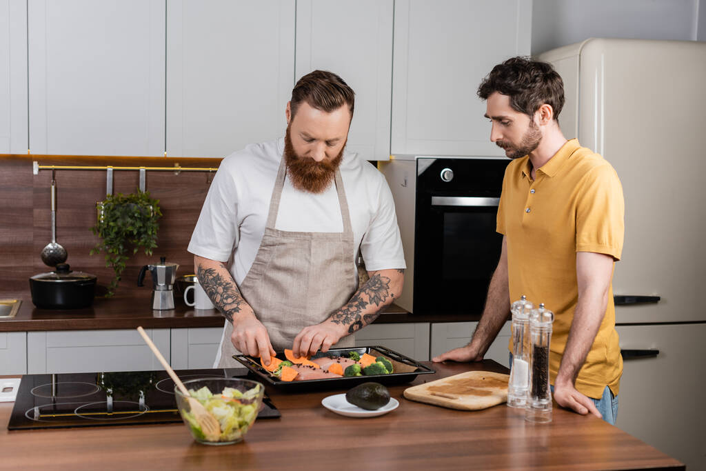 Gay man putting vegetables and chicken fillet on baking sheet near partner in kitchen  - Fotó, kép