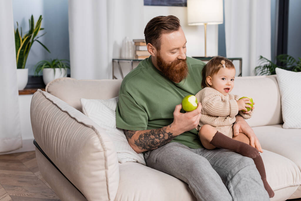 Hombre tatuado sosteniendo manzana e hija bebé en el sofá en casa  - Foto, imagen