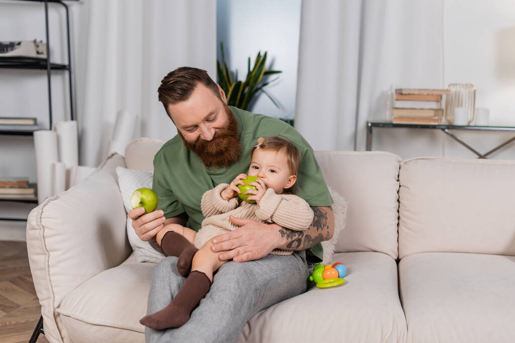 Padre barbudo positivo sosteniendo manzana y mirando a la hija del niño en el sofá  - Foto, Imagen