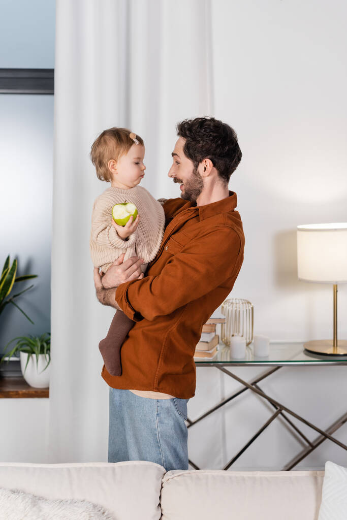 Vista lateral del padre alegre sosteniendo niña con manzana en la sala de estar  - Foto, imagen